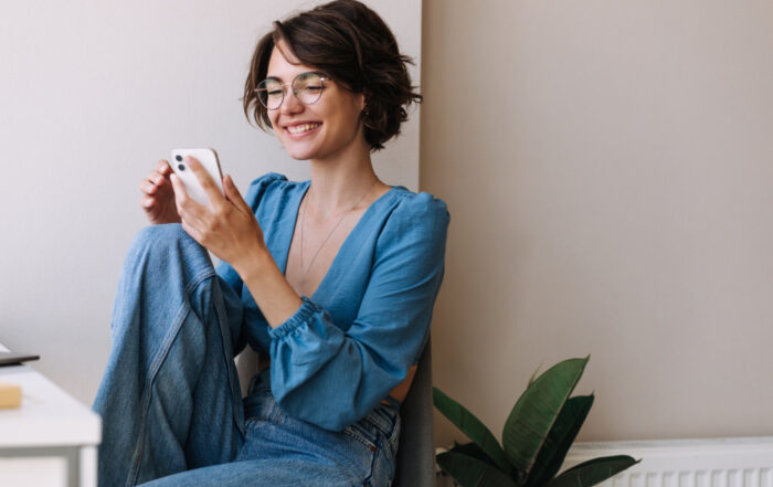 Woman in a blue blouse sitting against a wall, smiling at her phone. A potted plant is visible beside her.